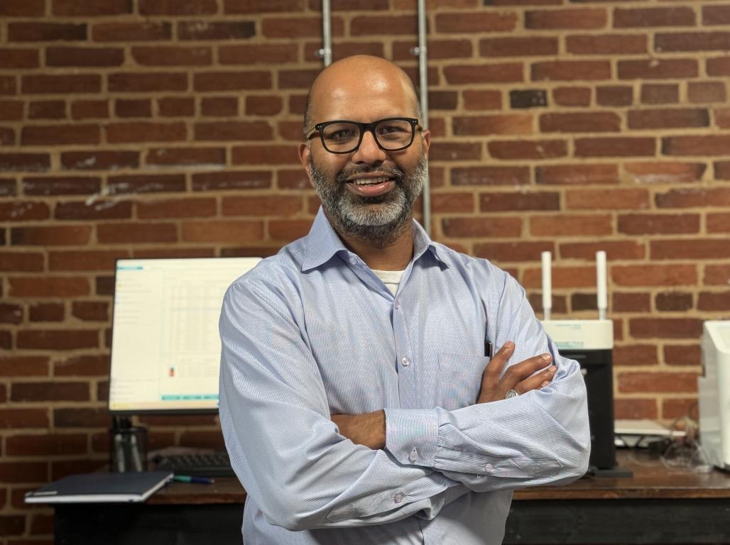 Smiling man with folded arms standing in an office in front of a brick wall and computer workstation.