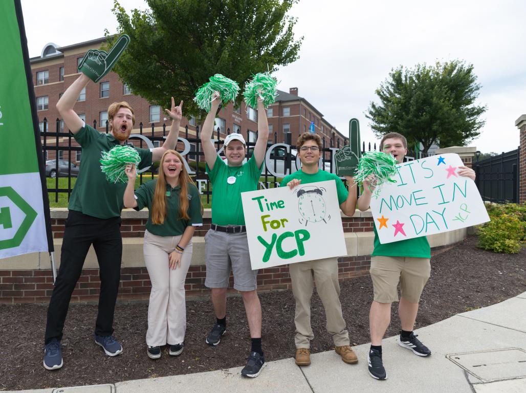 Five students in green shirts cheer with pom-poms and signs outside York College.
