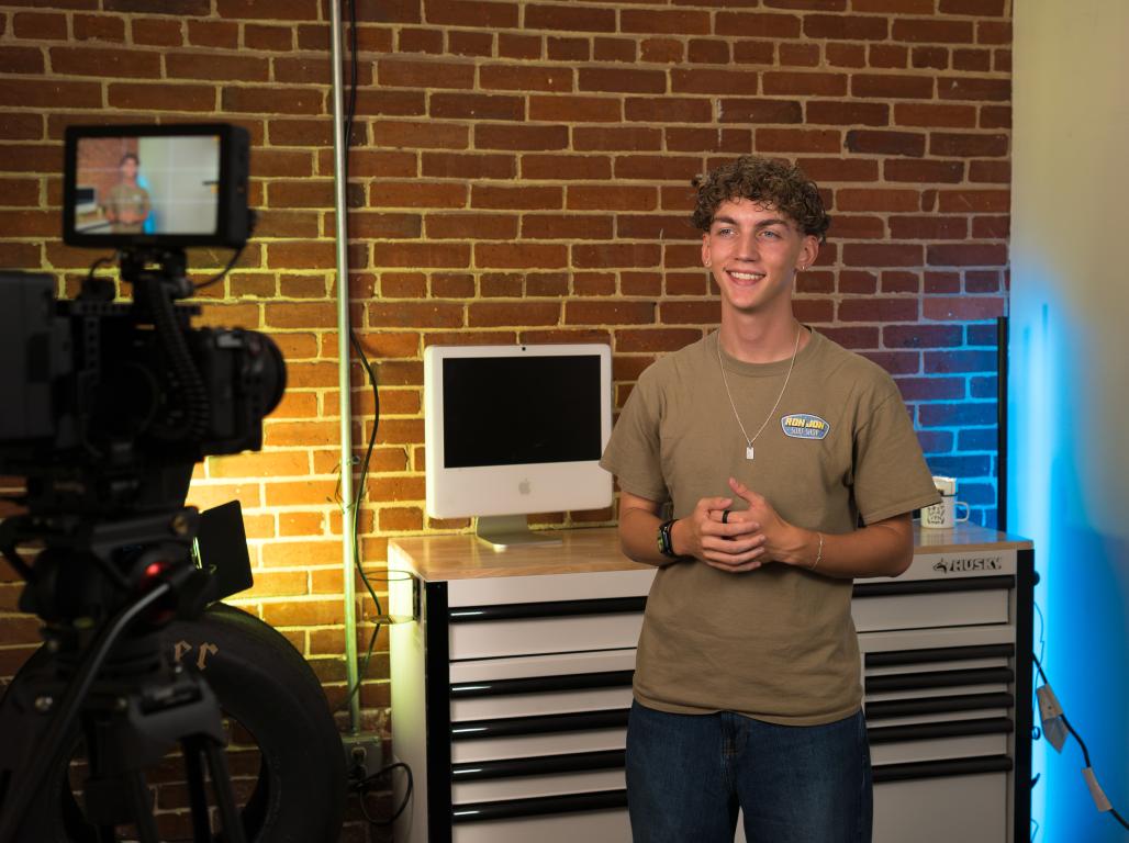 A student stands in a production workspace in front of a brick wall, with a camera, lighting equipment, and computer workstation set up around him.