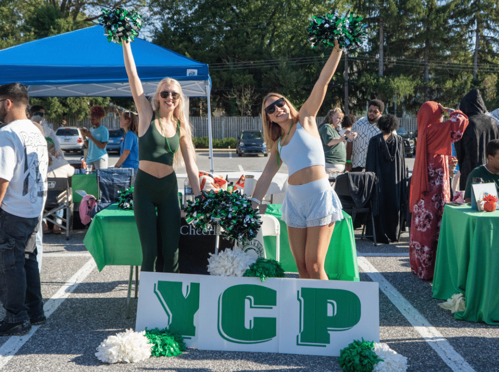 Two students hold green, black, and white pom-poms above a YCP sign during an outdoor campus event with student organization tables in the background.