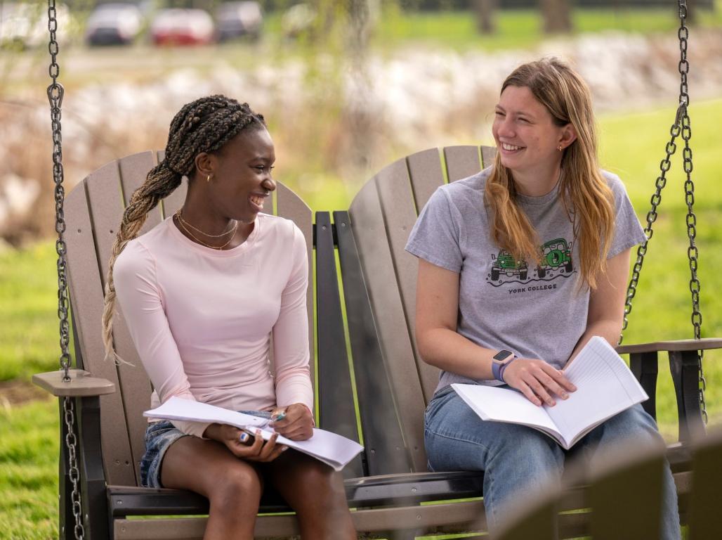 Two York College students smile while studying together on outdoor swing chairs.