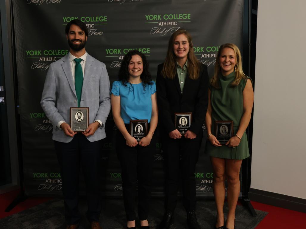 Four York College Athletic Hall of Fame inductees stand together holding commemorative plaques in front of a York College Athletic Hall of Fame backdrop.
