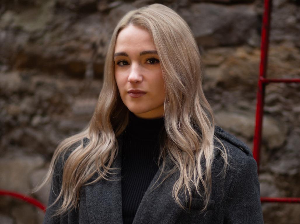 A woman wearing a dark coat and black turtleneck stands outdoors in front of a stone wall with red metal accents.