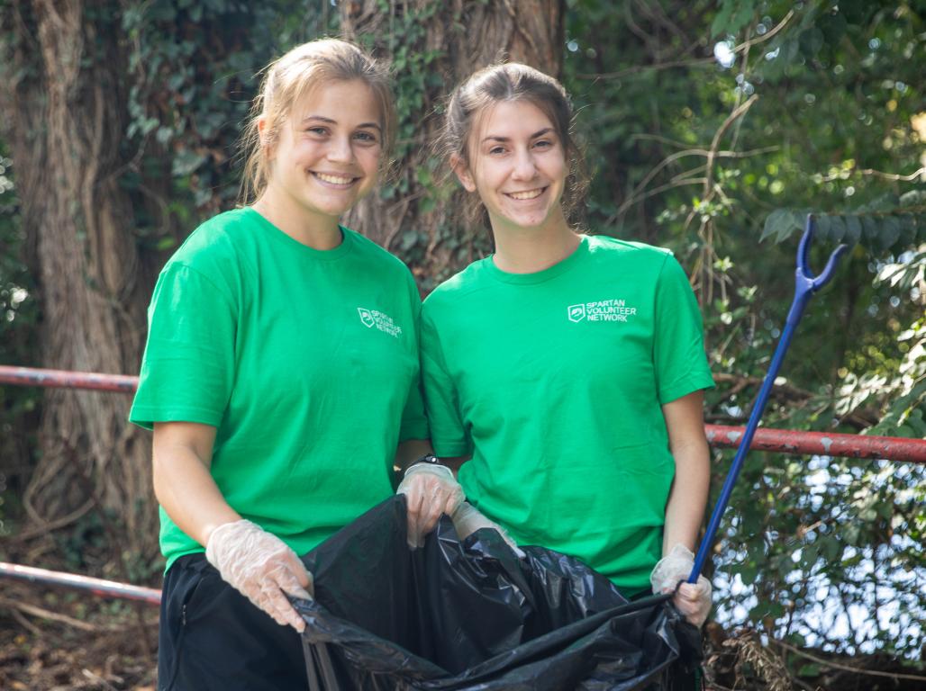 Two SpartanServe volunteers smile while holding a large trash bag and litter grabber during a community cleanup.