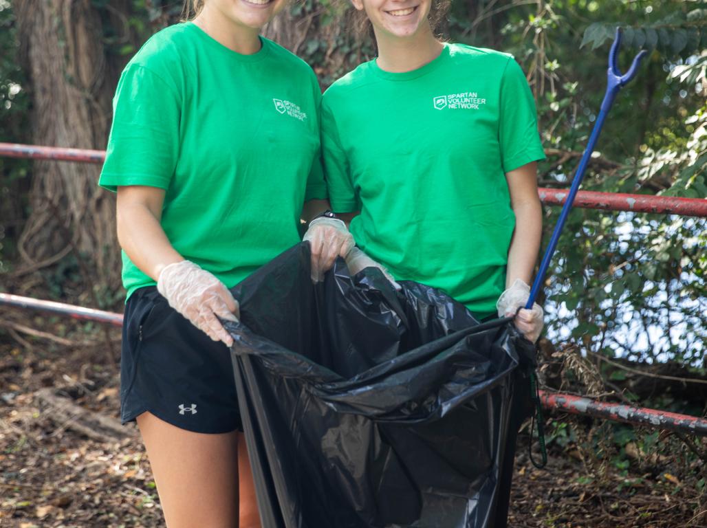 Two SpartanServe volunteers smile while holding a large trash bag and litter grabber during a community cleanup.