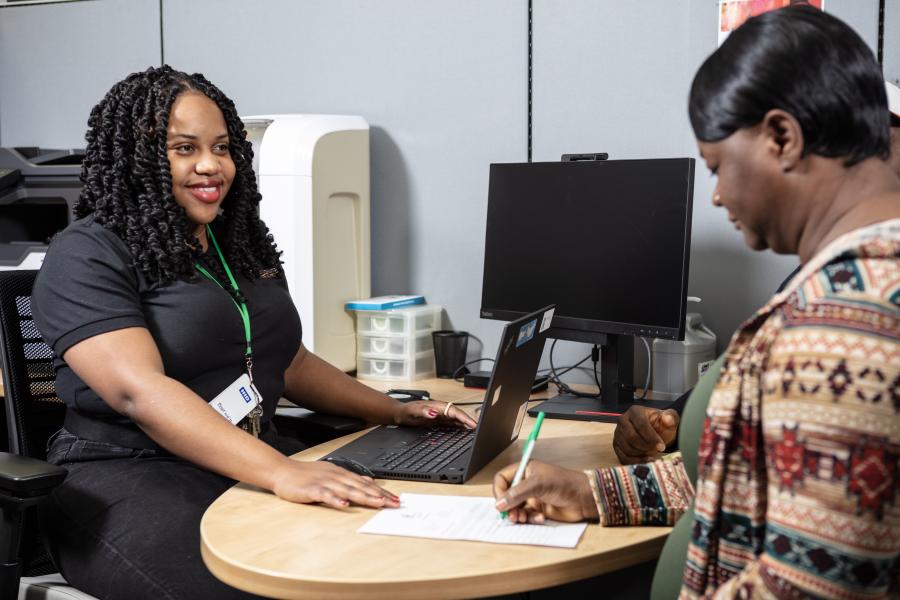 Person signing a paper at an office desk while another individual seated opposite smiles. There are computers and other office equipment visible.