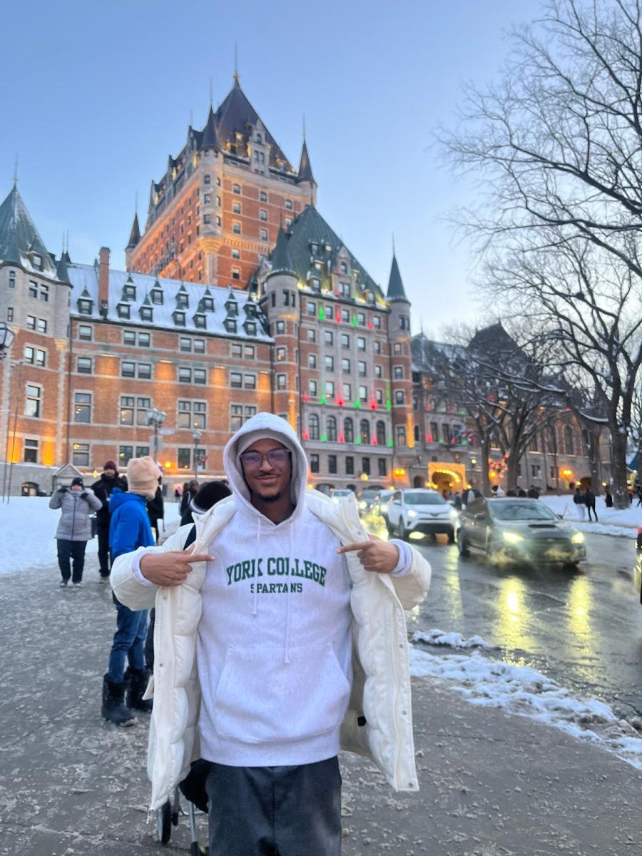 York College student standing in front of an ornate building on a winter day, pointing to the green lettering on their white hoodie, which reads "York College Spartans." 