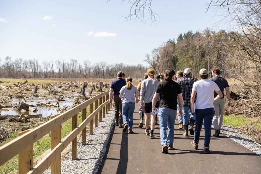 A group of people walk away from the camera outdoors on a sunny day. They're on a paved path with a wooden fence on one side and a muddy field visible beyond the fence.