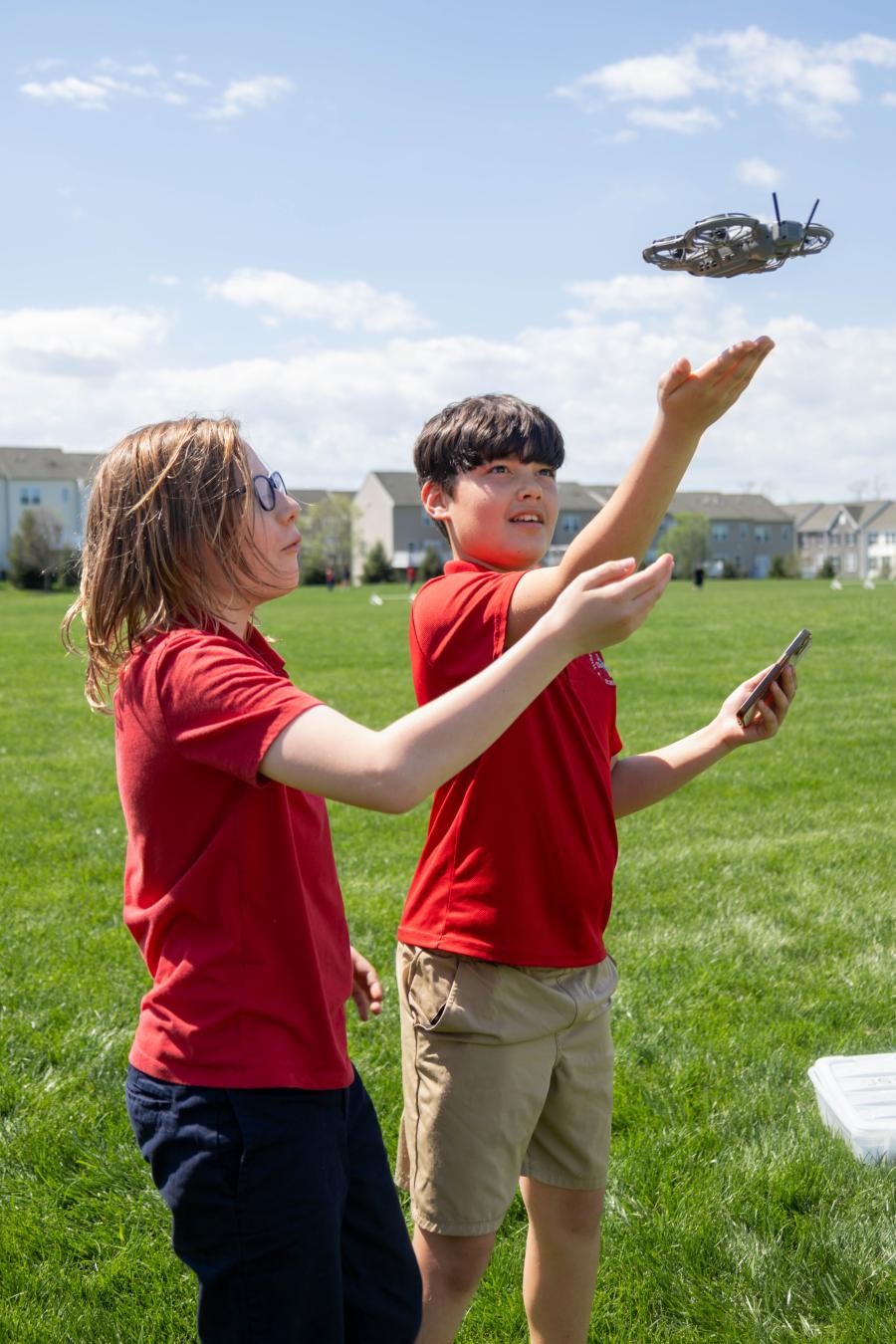 Two children wearing red shirts hold their hands up as a small drone takes flight on a sunny day.