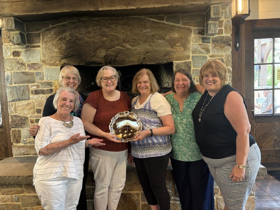 A group of people pose for a photo in front of a large, stone fireplace. The two in the center are holding a shiny, silver plate engraved with an award message.