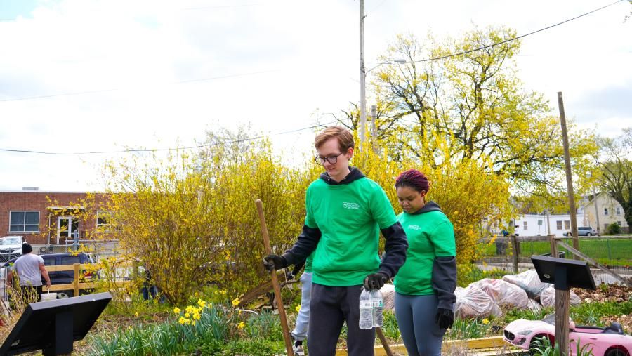 Two individuals wearing green tee shirts with "Spartan Volunteer Network" logos appear to be helping with a garden cleanup. They are both holding rakes and one is carrying bottled water.
