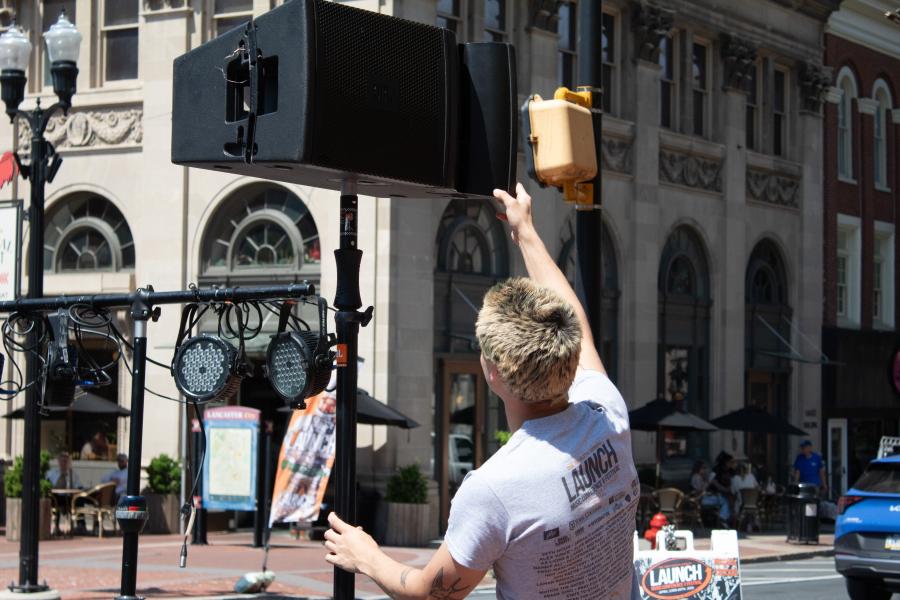 Person wearing a white tee shirt with a "LAUNCH Music Festival" logo on the back adjusts a speaker on a stand. The setting appears to be an outdoor city square.