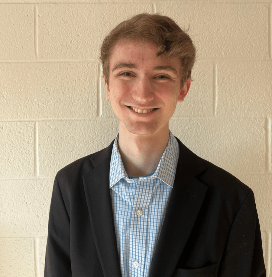 Christopher Magaha ’27 smiles for a professional headshot while wearing a dark blazer and blue checkered shirt against a light-colored brick wall.