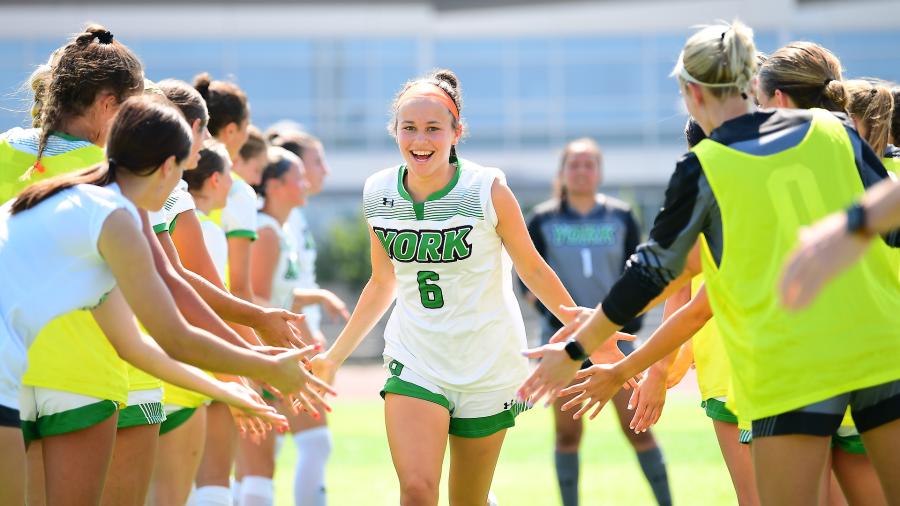 York College women’s soccer player wearing number 6 smiles as she runs through a tunnel of teammates offering high-fives on the field.