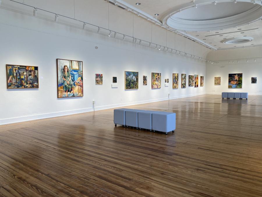 Gallery Hall at Marketview Arts with paintings displayed along white walls, polished wood floors, track lighting, and gray seating in the center of the room.
