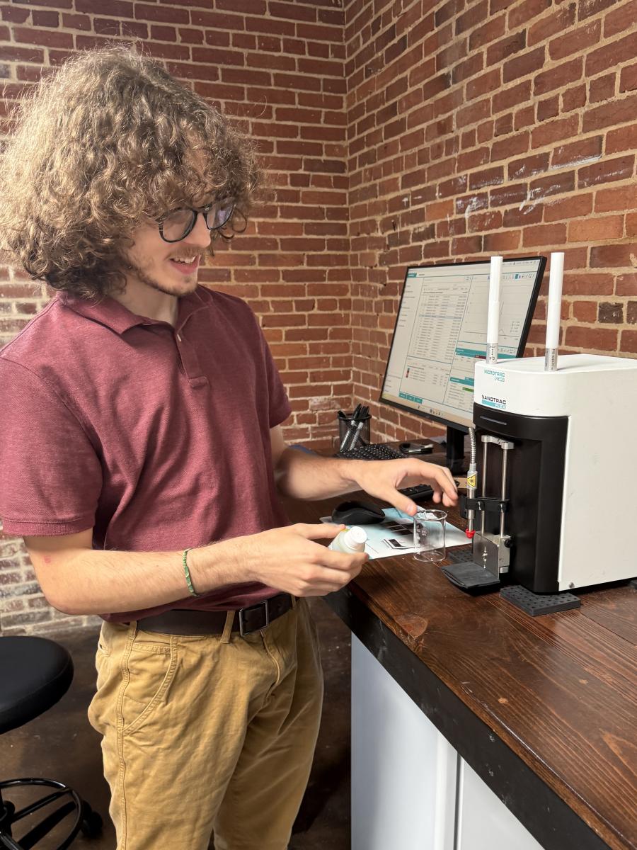 Person operating a laboratory instrument at a workstation with a computer in front of a brick wall.
