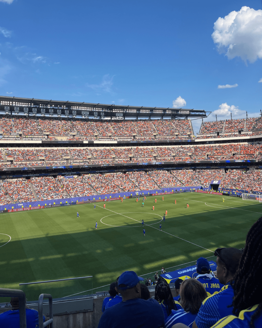 A wide view of a FIFA World Cup match shows players on the field and a packed stadium under a bright blue sky.