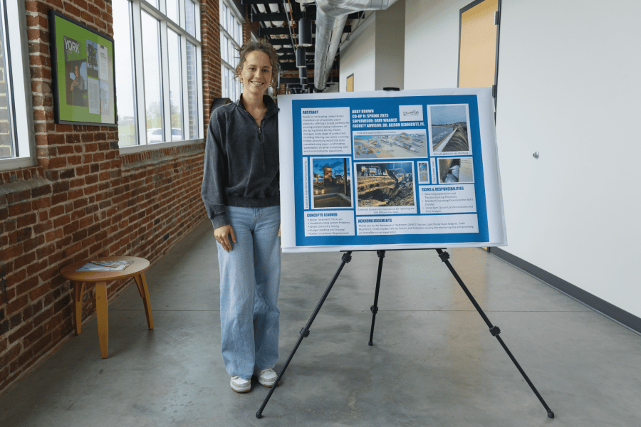 A York College Civil Engineering student stands beside a co-op presentation poster about her work with Pixelle Specialty Solutions in a brick-and-glass campus hallway.