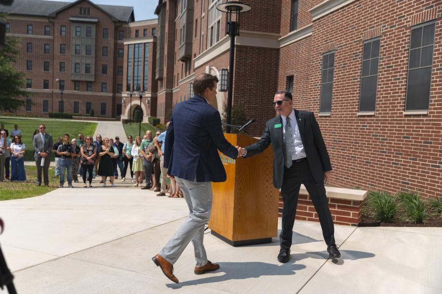 Two men shake hands beside a podium during an outdoor York College event, with attendees gathered nearby.