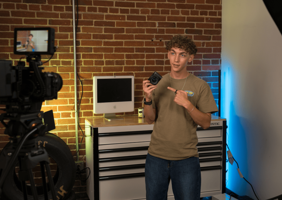A student stands in a production workspace holding a camera, with video equipment, lighting, and a computer workstation set up around him.