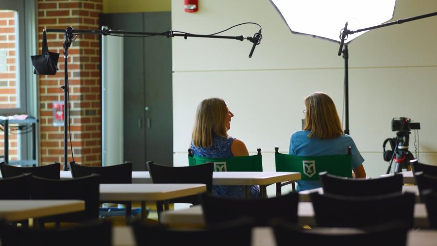 Two Lifelong Learning Initiative participants sit in green York College chairs during a filmed interview, surrounded by cameras, microphones, and lighting equipment.