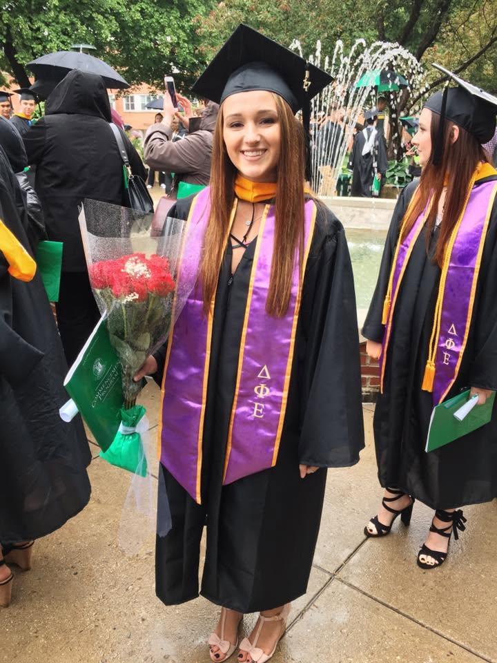 A York College graduate smiles in cap and gown while holding a bouquet of flowers near the campus fountain, with other graduates gathered in the background.