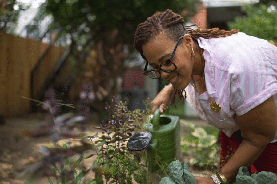 A woman smiles while watering plants in an outdoor community garden.
