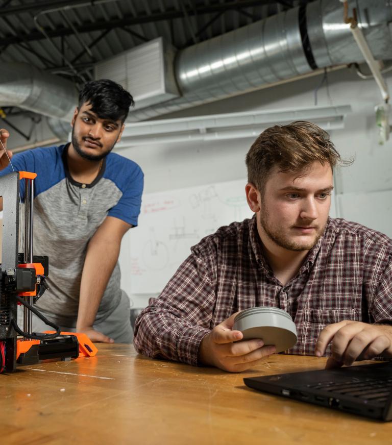 A student works with the filament of a 3D printer while a second student looks at an open laptop.