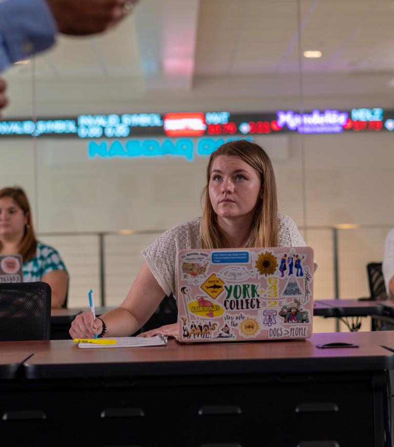 Students listen attentively to the professor teach while also taking notes in the NASDAQ Training lab