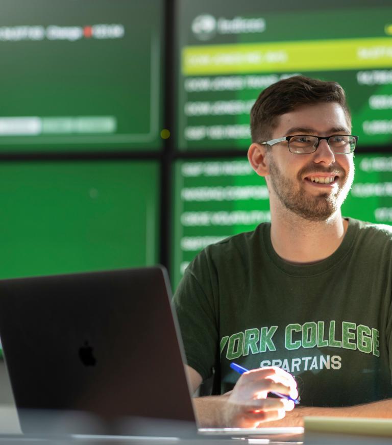 A student sits with a laptop holding a pen over a notepad in front of a large video display of market information.