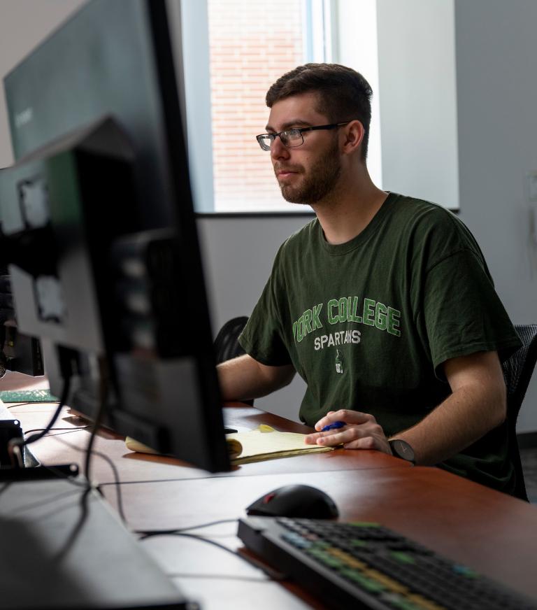 A student sits at a desk with two large monitors, a mouse, and keyboard. An interactive screen displaying business data is visible on the wall behind him.