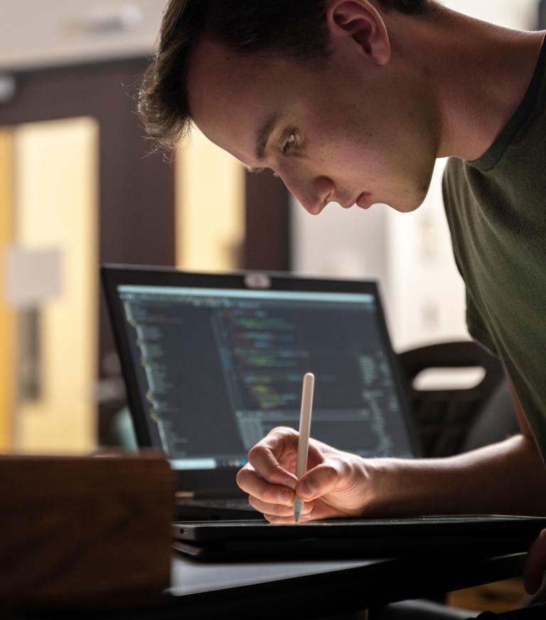 A student writing on a piece of paper with computer science software on the laptop behind them.