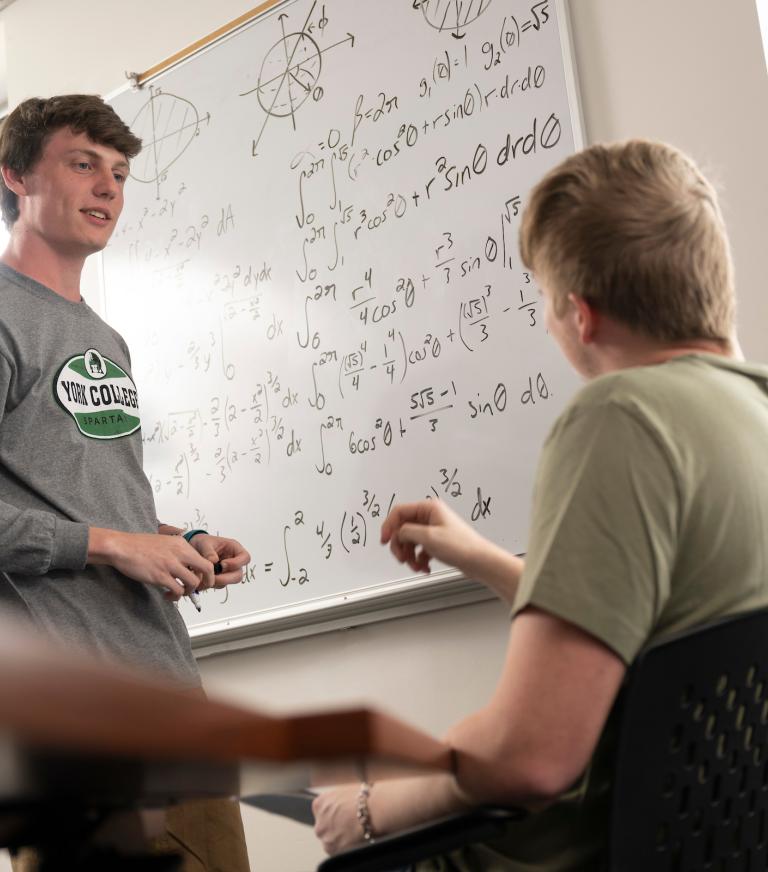 Two Students discuss a math problem written on a whiteboard 