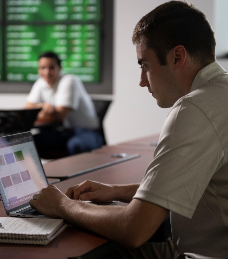 A student works on a laptop with an open notebook to the left.