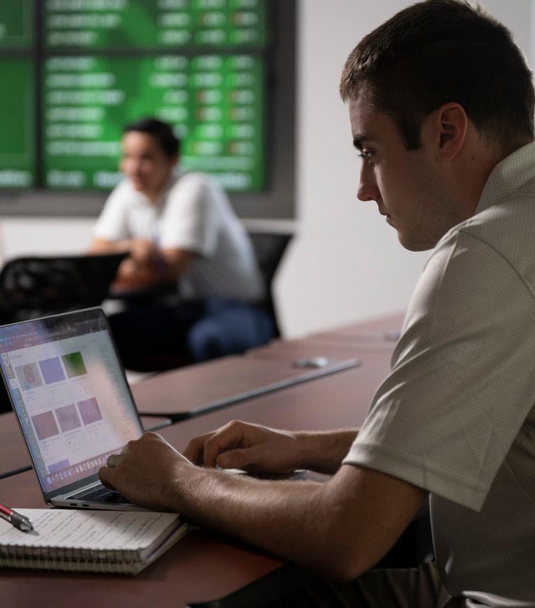 A student works on a laptop with an open notebook next to it.