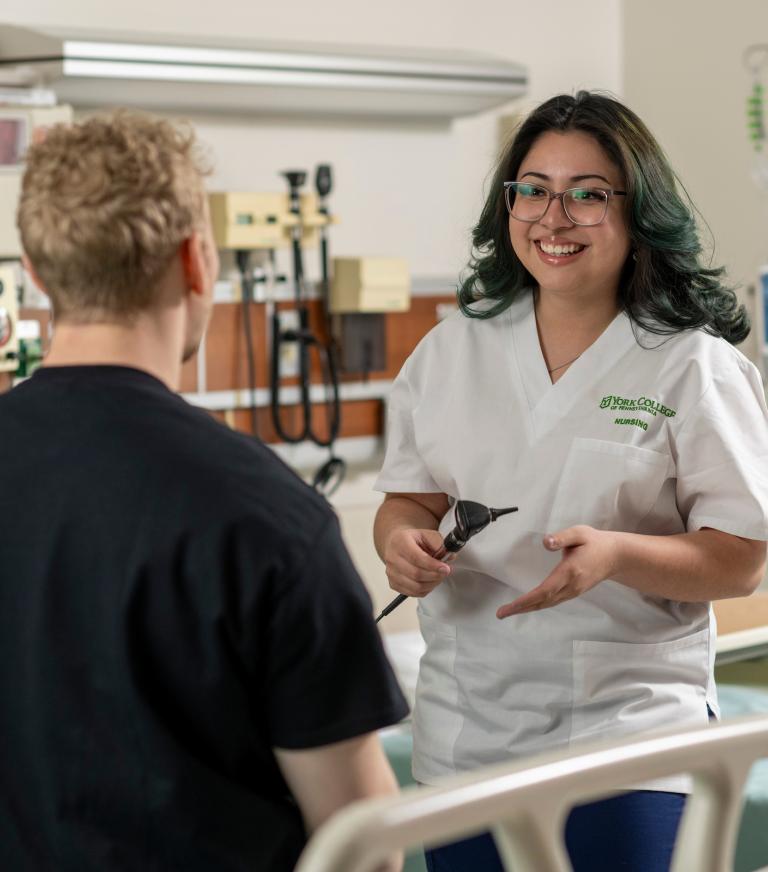 A nursing student smiles at a patient on an exam table in a clinical environment.