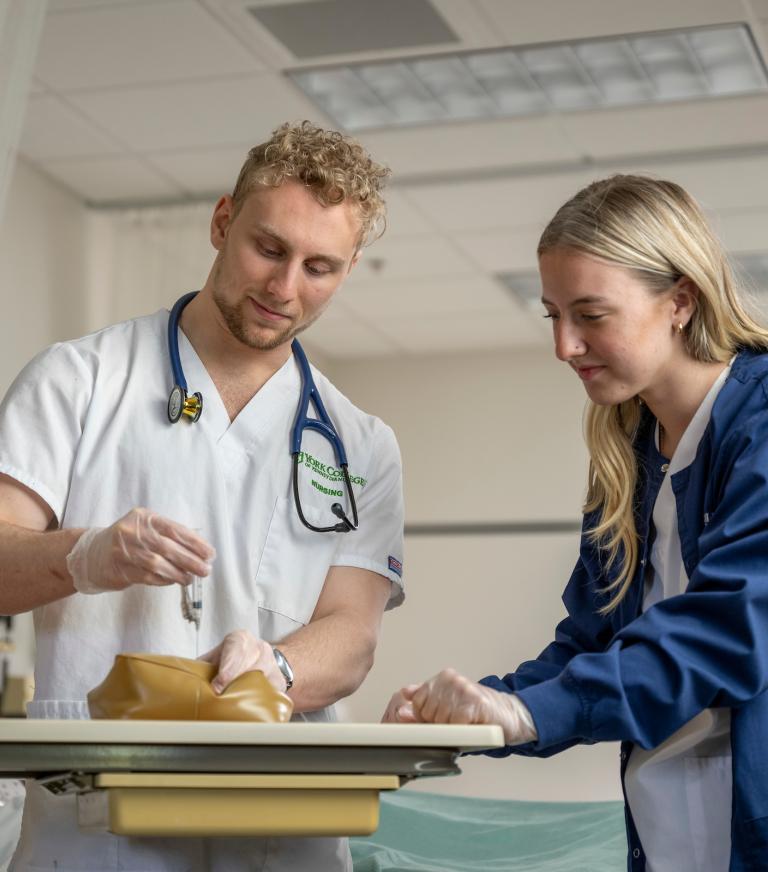 Two students in the nursing lab practicing giving a shot.