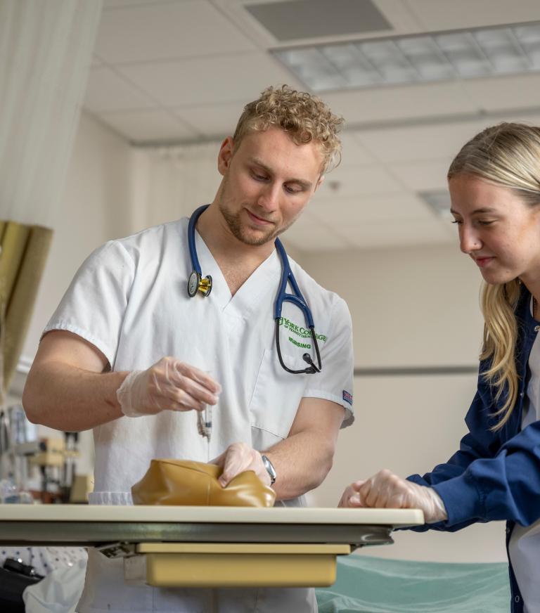 Two students in the nursing lab practicing giving a shot.