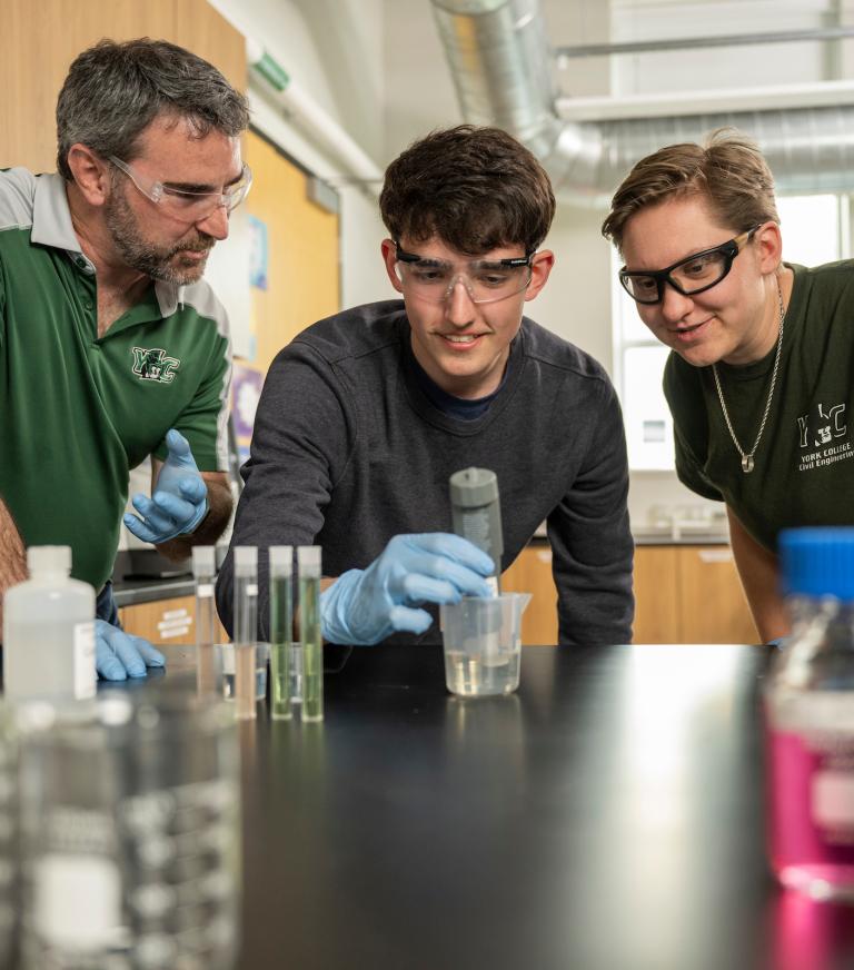 An instructor and a student observe a student holding a pipette in a beaker of solution.