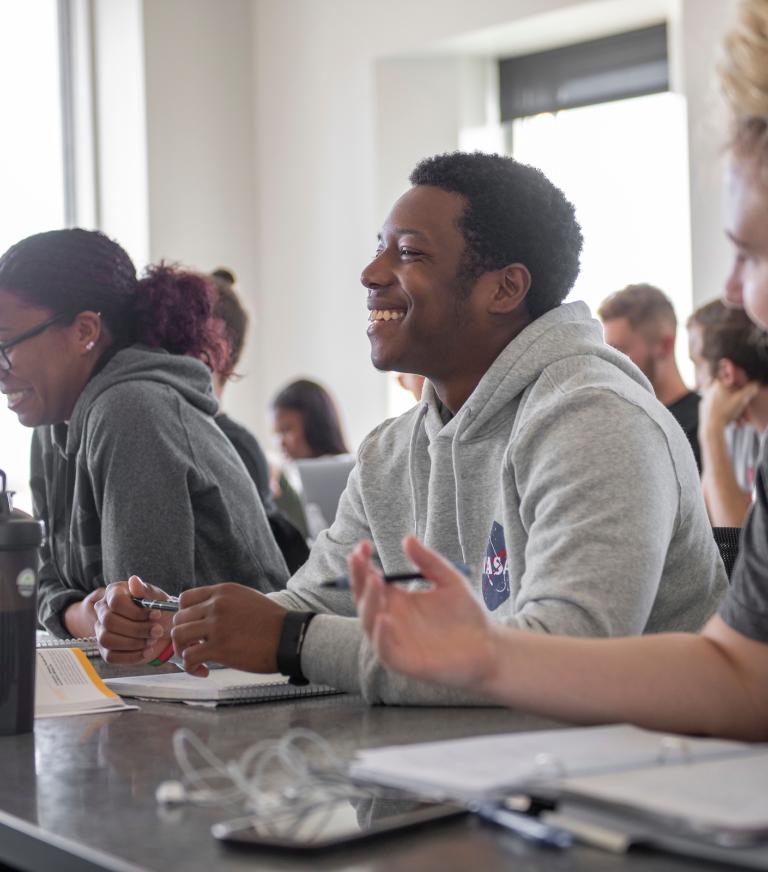 Three students sit at a classroom table smiling with open notebooks during a class.