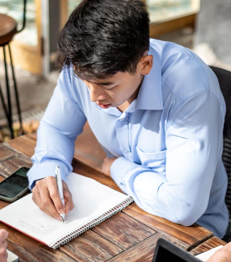 Two students work at a table looking at a notebook. One student writes in the notebook. 