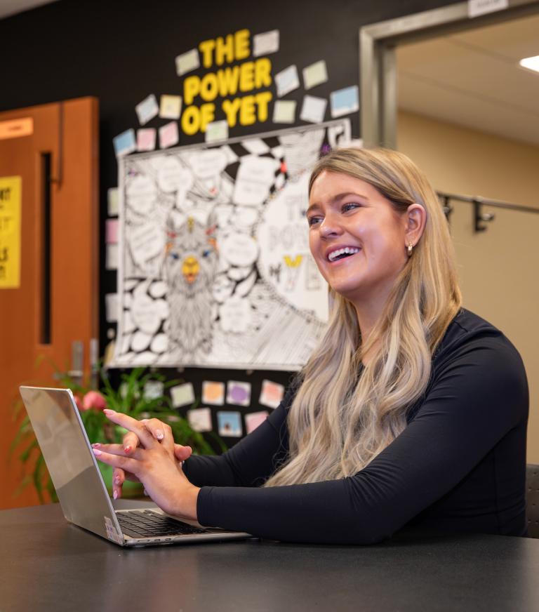 A student smiles in the Writing Center, laptop on the desk in front of her. A decorated bulletin board is visible in the background.