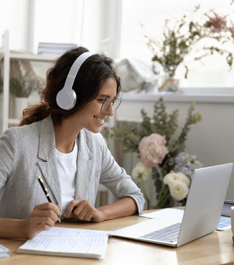 An adult student wears headphones and takes notes as she looks at a laptop.