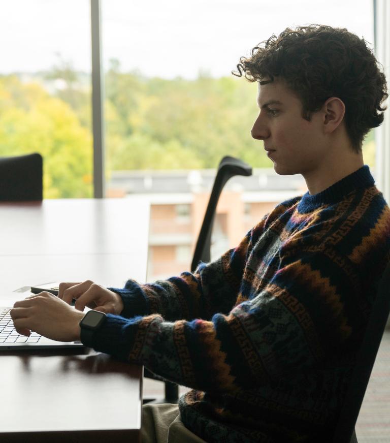 A student types at a laptop, his desk positioned in front of a large window letting in natural light.