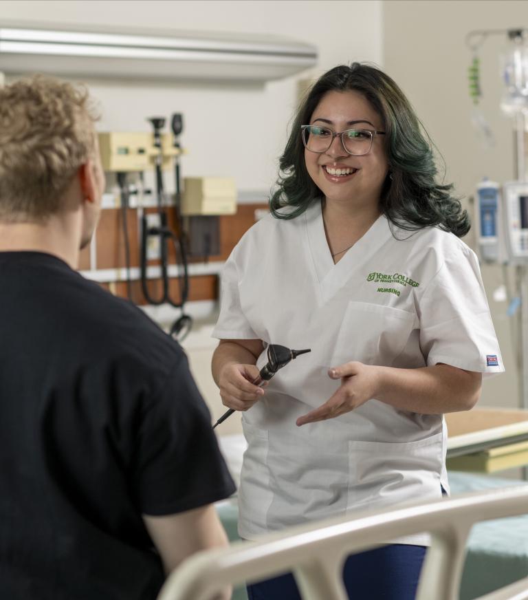 Smiling York College Accelerated BSN student faces another student to practice gathering vitals in a hospital simulation lab