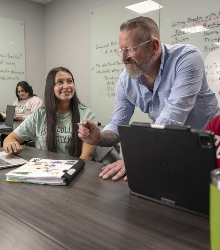 Instructor assisting students in a collaborative classroom with laptops, while whiteboards display notes on democracy and voting systems.