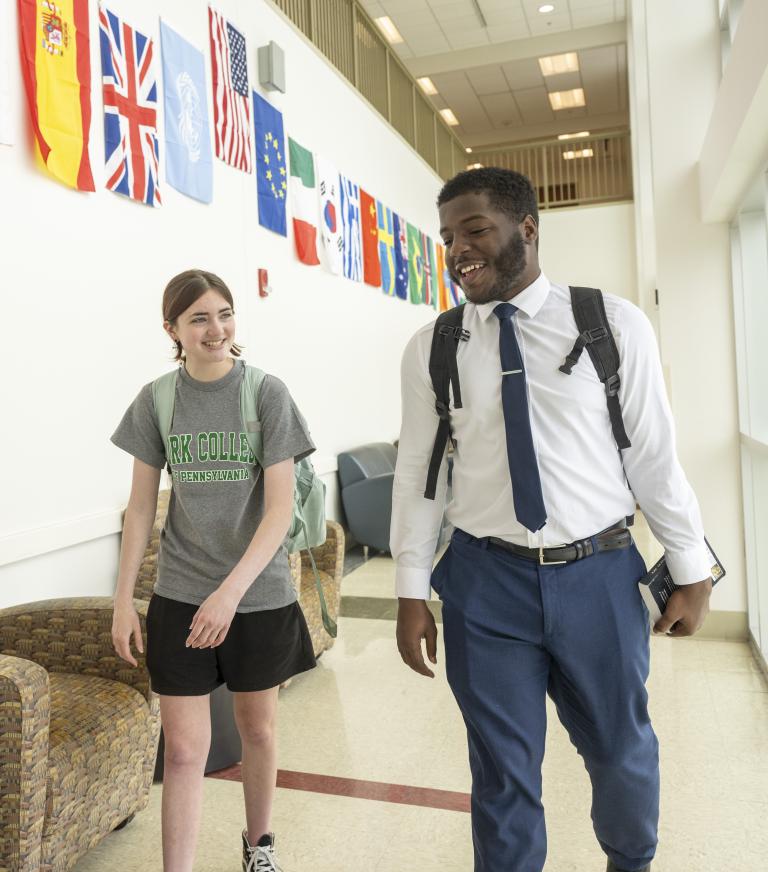 Two students walk through a hallway lined with international flags, smiling as they talk on a college campus.