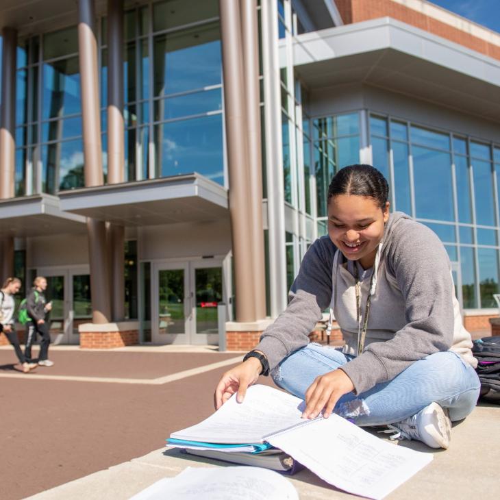 Student reads notebook while sitting on ledge with glass building in background