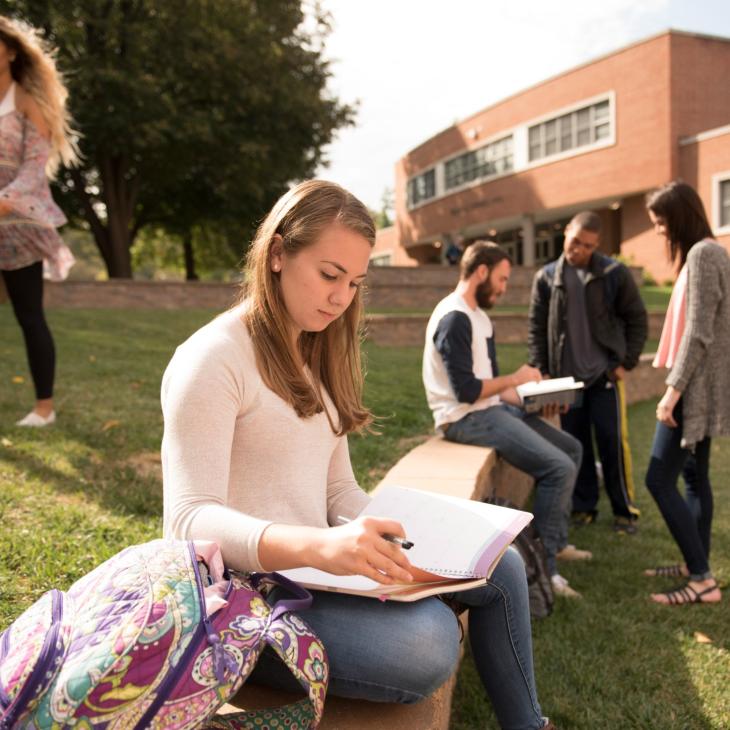 Student writes in notebook while sitting on bench on campus mall