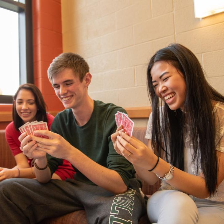 Four students sitting on a couch laughing and playing a game of cards.